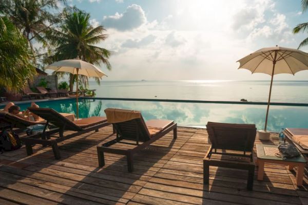 An infinity pool overlooking the ocean is surrounded by lounge chairs and umbrellas, with a few people relaxing, set against a beautiful sky.