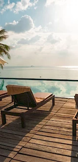 Lounge chairs, umbrellas, and a pool deck overlook the ocean with a sunny sky and palm trees, creating a serene tropical setting.