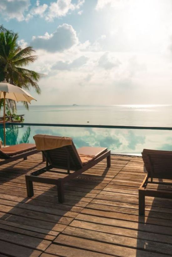 Poolside scene with lounge chairs, umbrellas, a wooden deck, and a serene view of the ocean under a partly cloudy sky.