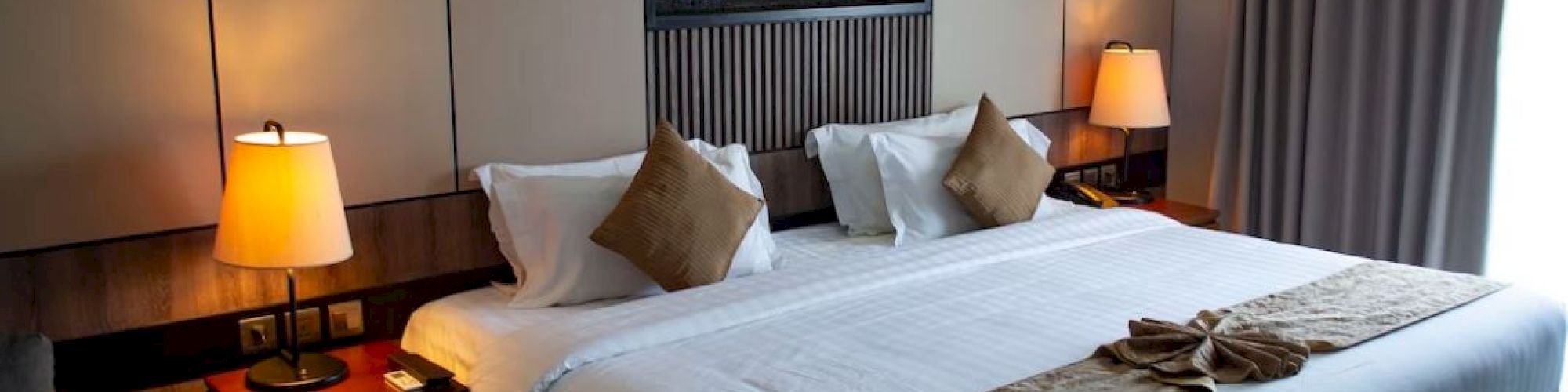 This image shows a cozy hotel room with a large bed, two side tables with lamps, a decorative wall feature, and a window with heavy curtains.