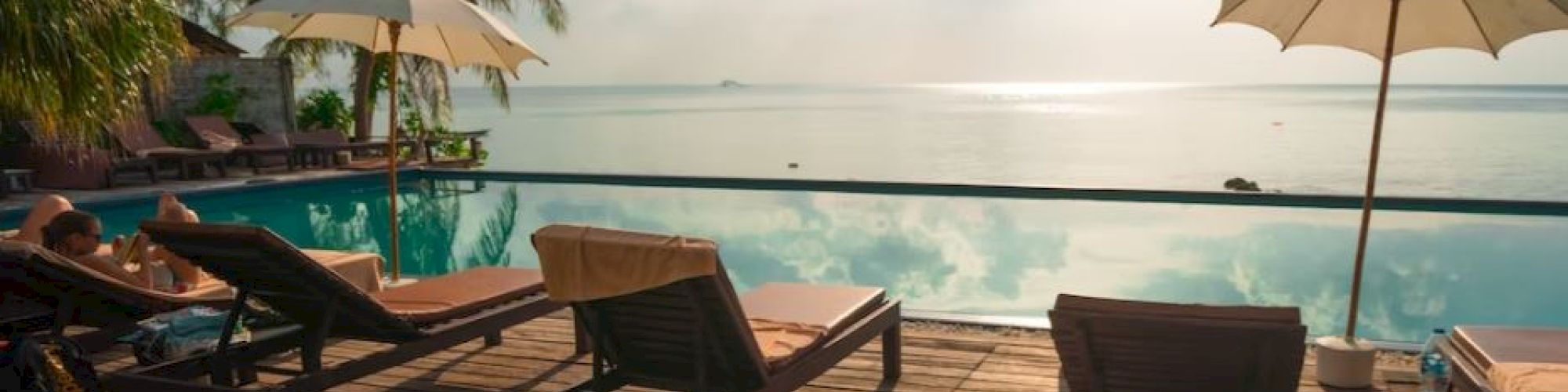 A serene poolside scene featuring lounge chairs and umbrellas on a wooden deck, with a calm ocean and a bright sky in the background.