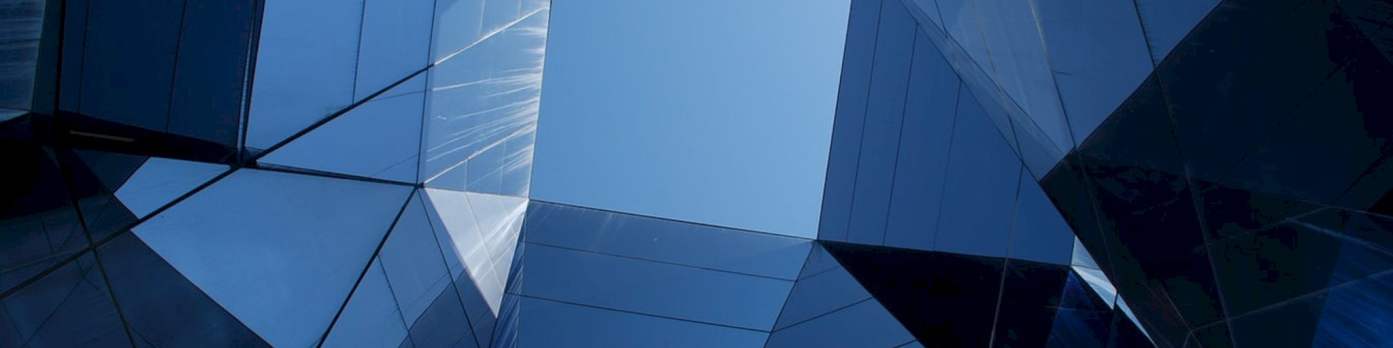 The image shows a geometric arrangement of mirrored glass panels creating a polygonal structure with a central open space revealing the sky.