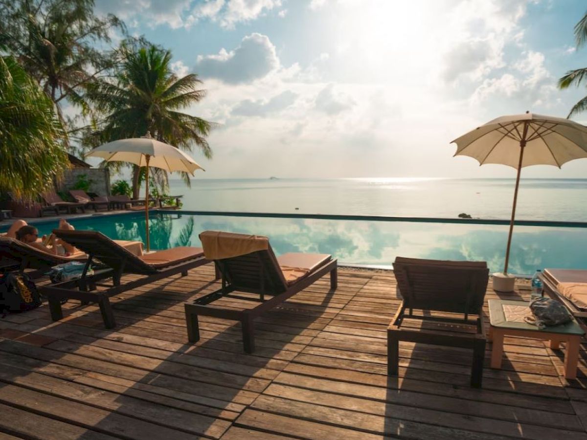 A serene poolside scene with lounge chairs, umbrellas, and a wooden deck overlooking a calm body of water, set against a backdrop of palm trees.