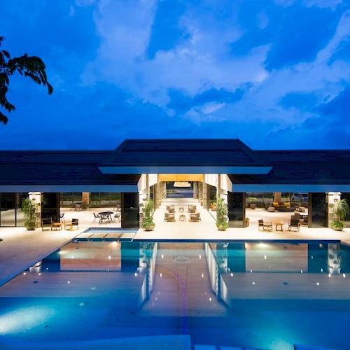 The image shows a modern, luxurious building at twilight with a large, well-lit swimming pool in front, surrounded by lounge chairs and greenery.