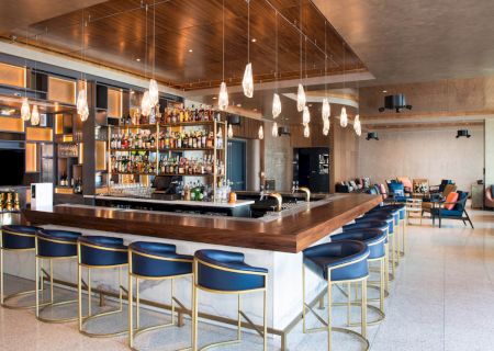 A modern bar with a fully stocked counter, blue bar stools, hanging pendant lights, and a lounge area with seating in the background.