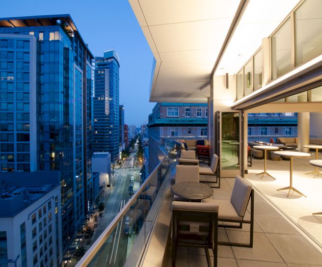 A modern balcony at dusk overlooks a city street with tall buildings, featuring outdoor seating and tables, and large windows illuminating the space.