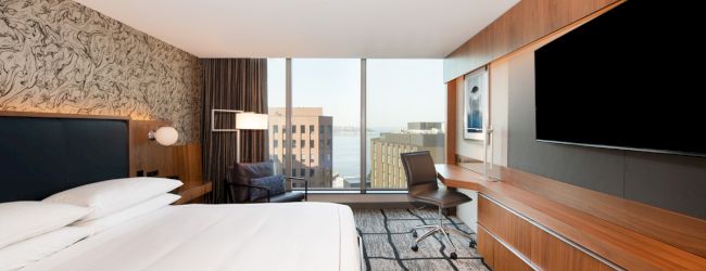 A modern hotel room features a large bed, desk, and chair with cityscape and water views through floor-to-ceiling windows.