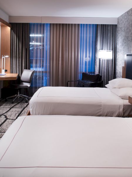 A modern hotel room with two beds, a TV, desk, chair, and stylish decor, featuring patterned wallpaper and large windows with curtains.