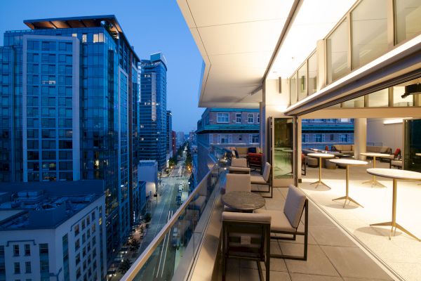 A modern rooftop terrace at dusk overlooking a cityscape with tall buildings and a street below, featuring seating and tables.