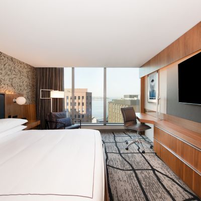 A modern hotel room with a large bed, wall-mounted TV, desk, chair, and a view of city buildings through floor-to-ceiling windows.