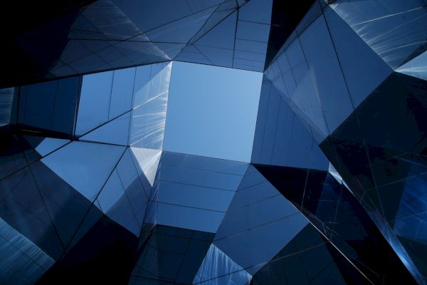 The image shows an upward perspective of modern glass buildings with reflective surfaces, creating abstract geometric patterns against a blue sky.