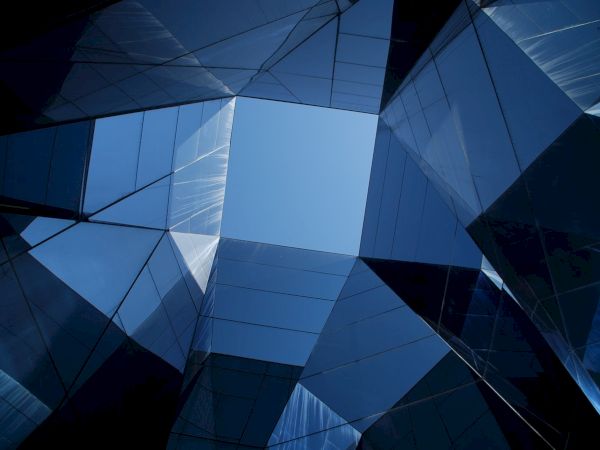 The image shows an upward perspective of modern glass buildings with reflective surfaces, creating abstract geometric patterns against a blue sky.