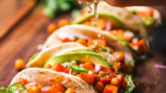 The image shows a hand squeezing a lime over a row of tacos filled with chickpeas, diced vegetables, lettuce, and garnished with cilantro.