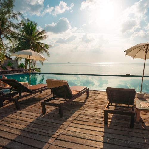 The image shows a serene poolside with lounge chairs, umbrellas, palm trees, and a stunning view of the horizon over the water, under a partly cloudy sky.