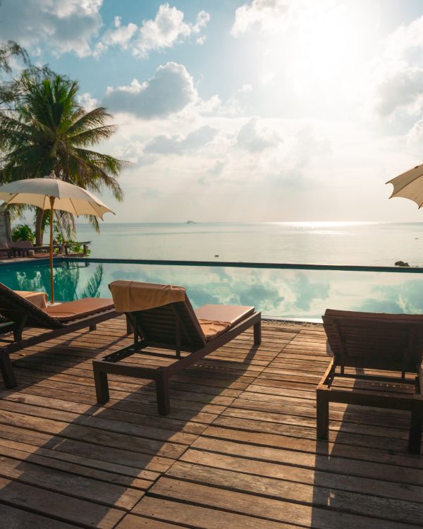 The image shows a serene poolside with lounge chairs, umbrellas, palm trees, and a stunning view of the horizon over the water, under a partly cloudy sky.