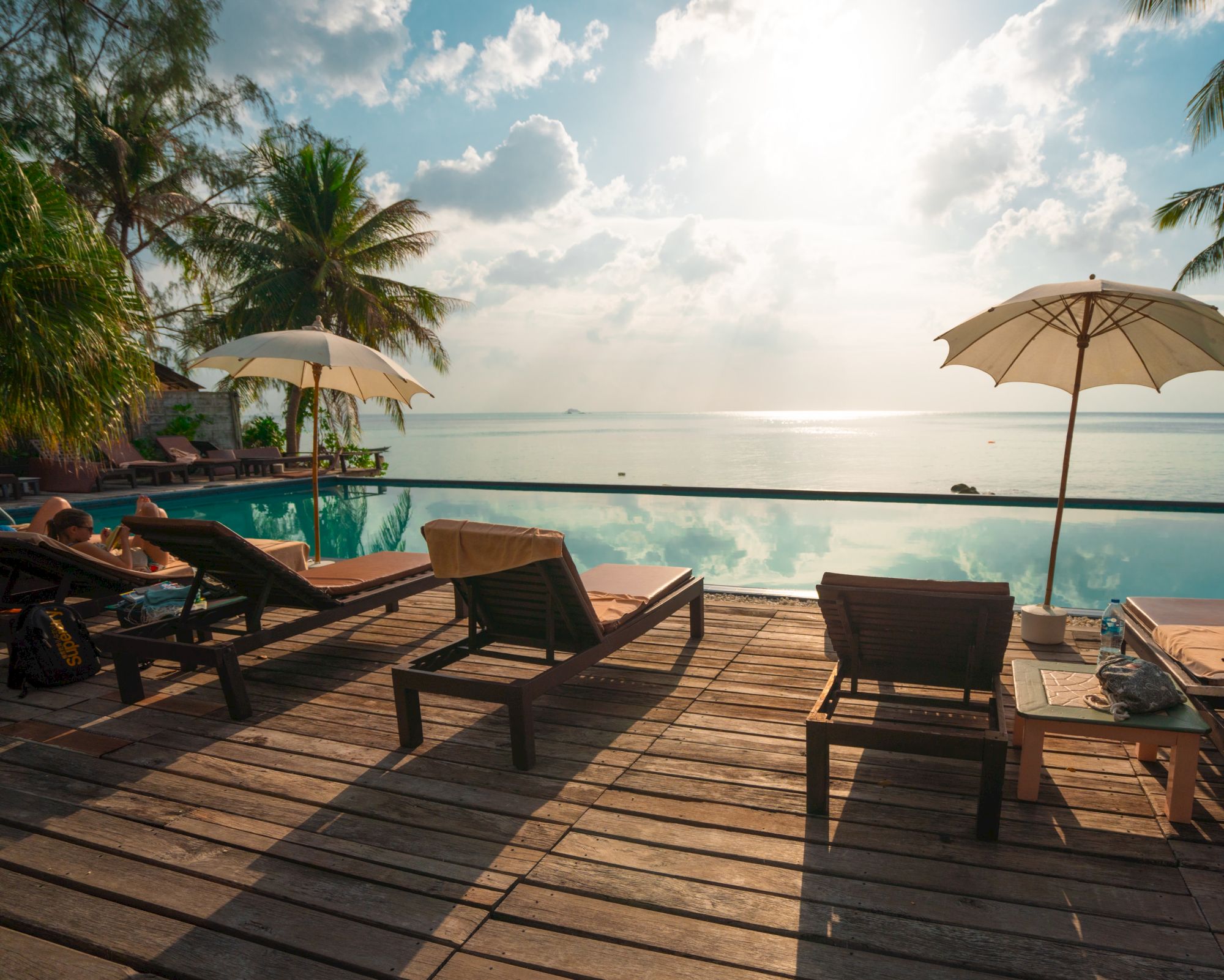 Wooden deck with lounge chairs, two umbrellas, and a gorgeous ocean view under a sunny sky dotted with clouds.