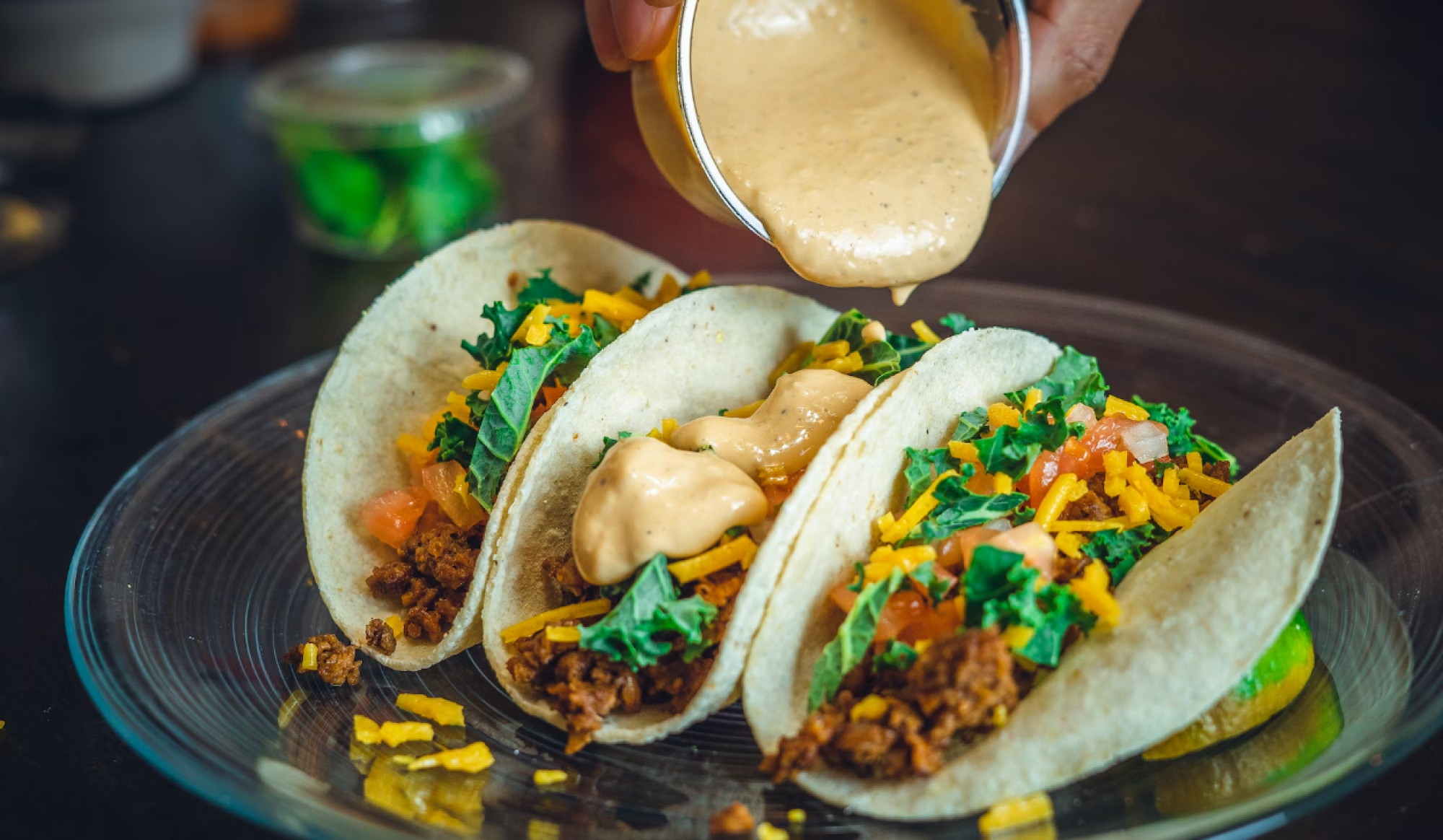 The image shows three tacos on a plate with someone pouring creamy sauce over them. The tacos are filled with meat, cheese, and greens.