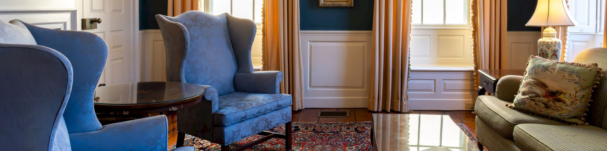 A beautifully furnished living room features blue armchairs, a beige sofa, framed art, and large windows with yellow curtains, creating a cozy atmosphere.