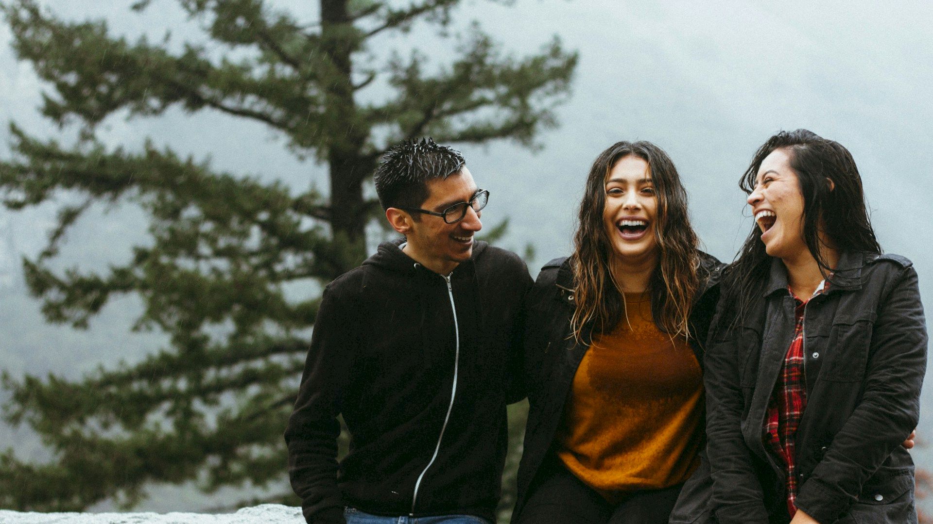 Three people sit on a stone ledge, laughing and enjoying themselves, with a blurred Newport, RI scenic forest background.