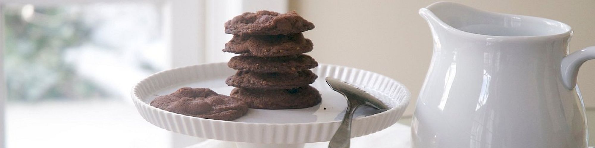 A stack of chocolate cookies on a cake stand, a white pitcher, and a floral-embroidered cloth on a table by the window.