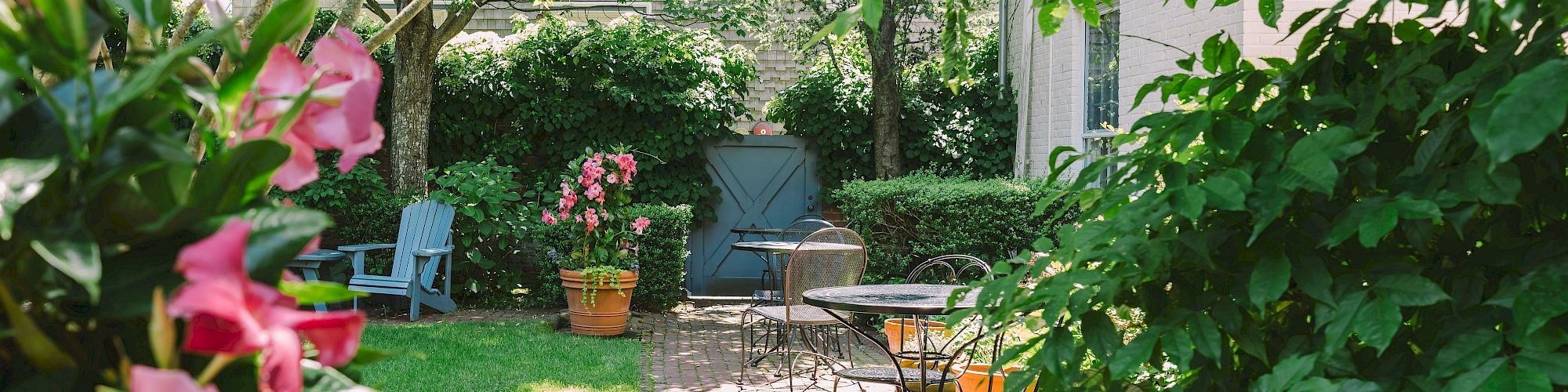 A lush garden with pink flowers, a brick path, outdoor seating, and a small blue door in the background, surrounded by greenery.