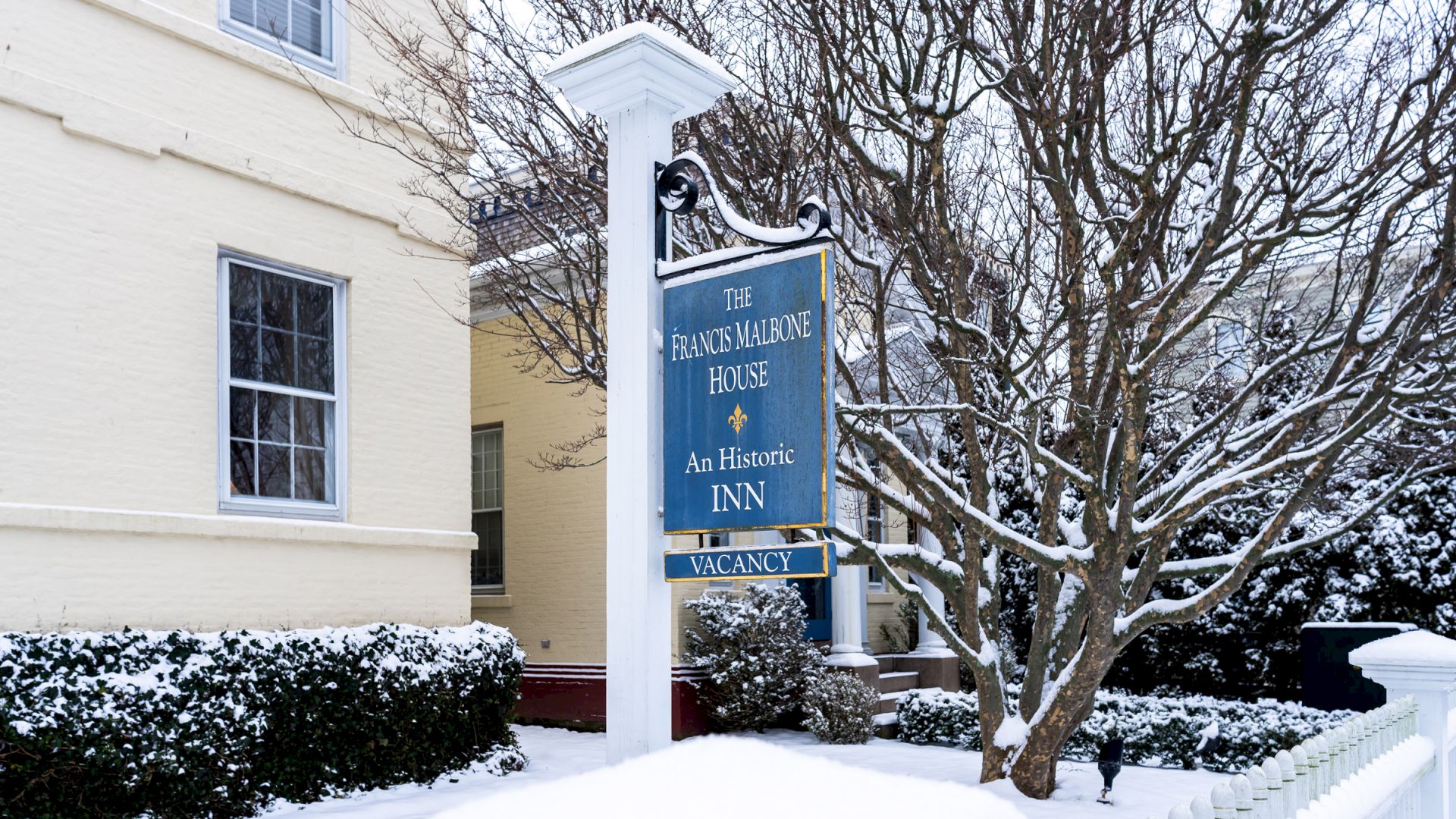 A blue sign on a post snow-covered reading "The Francis Malbone House. An Historic Inn. Vacancy.", outside a Newport, RI, hotel.