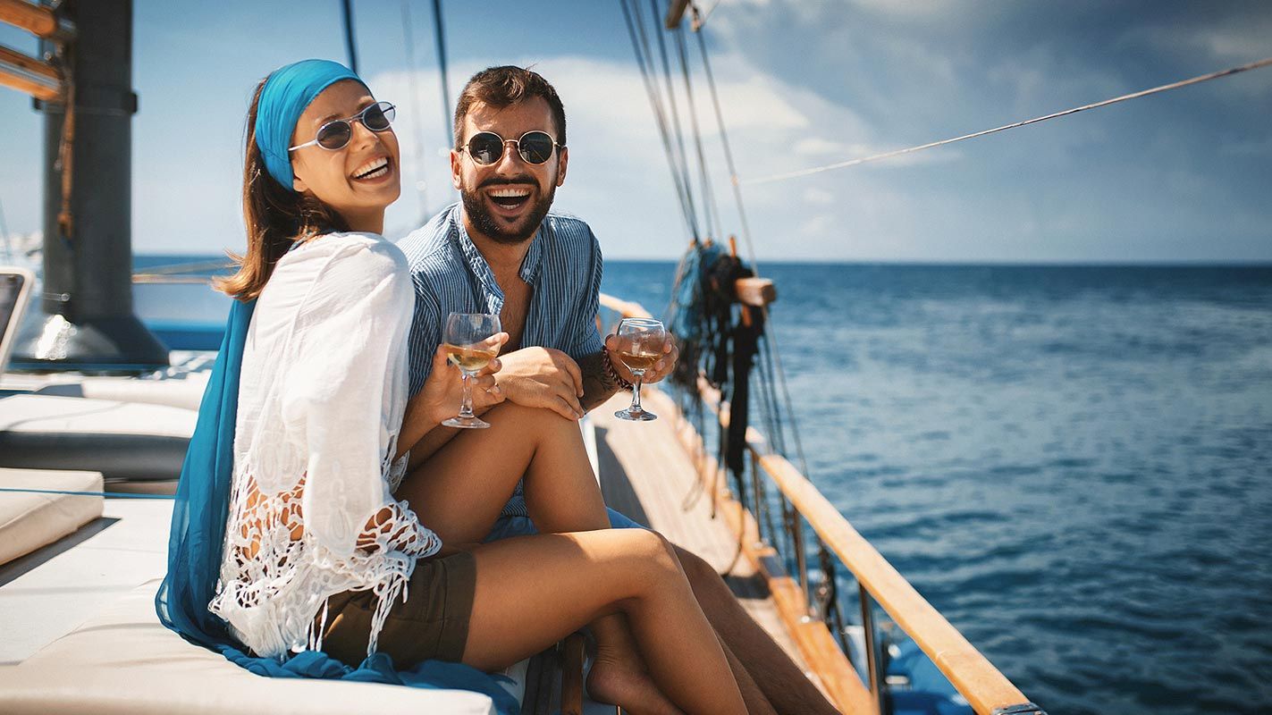 A smiling couple wearing sunglasses enjoying drinks and sitting on a boat on the ocean on a sunny day.