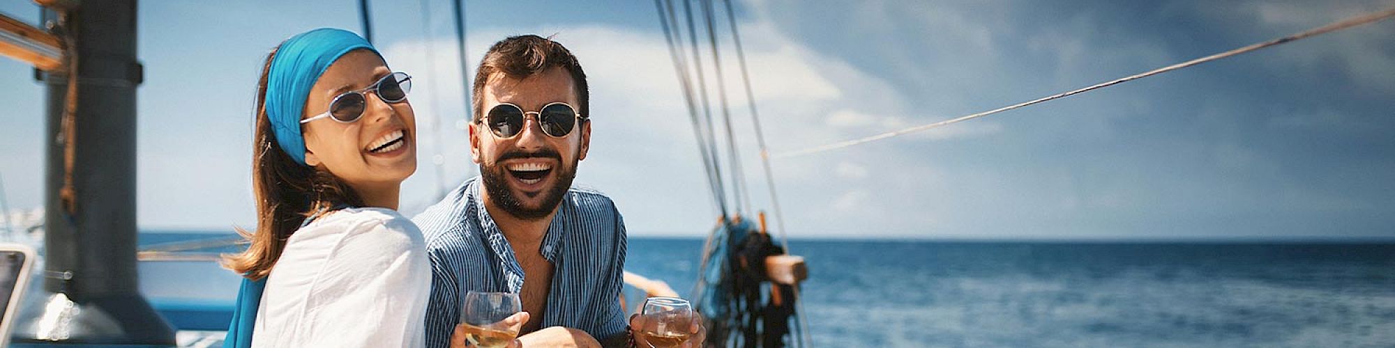 A smiling couple is sitting on a boat, enjoying drinks with the ocean in the background. Both wear sunglasses, enjoying a sunny day on the water.