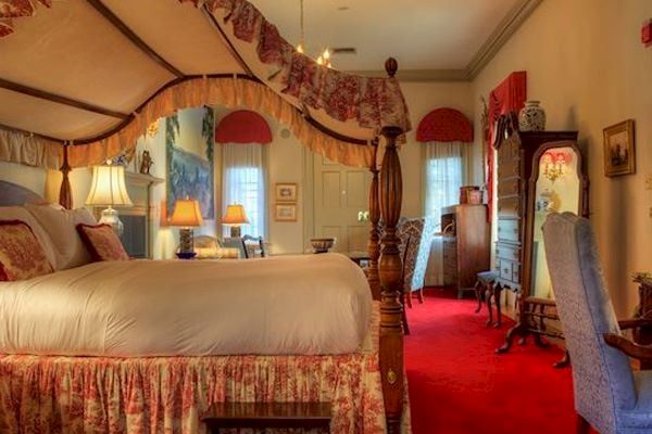 A well-furnished traditional bedroom with a four-poster bed, armchair, dresser, vanity, lamps, and a red carpet on the floor ending the sentence.