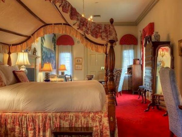 A cozy Newport, RI, guest room with a king-size canopy bed, a full-length mirror, lamps, and several armchairs.