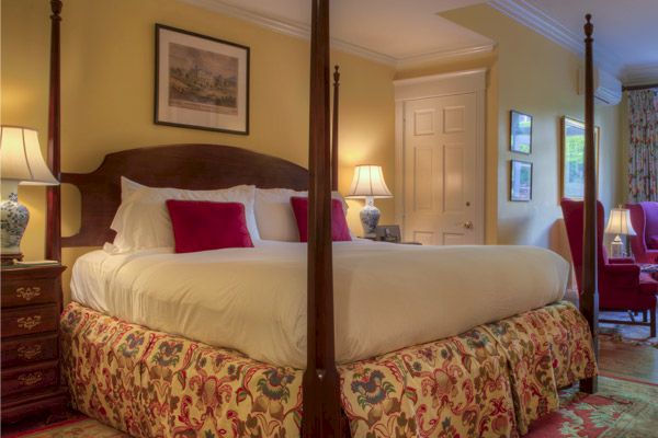 A cozy bedroom with a large bed, two side tables with lamps, a door, framed art, and a patterned bed skirt, upholstered furniture, and curtains.