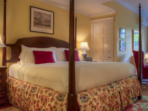 A cozy Newport, RI, guest room with a four-poster king-size bed, lamps, red armchairs, and paintings hung on the walls.