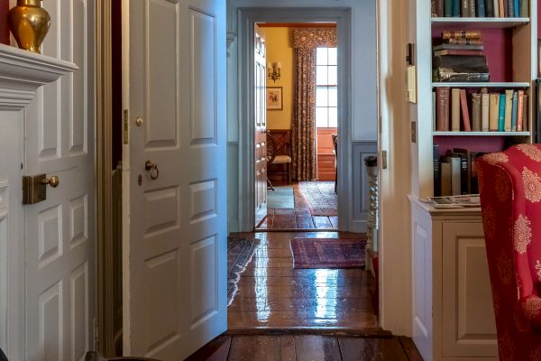 The image shows a cozy, well-furnished hallway with wooden floors, white doors, a bookshelf, and warm lighting, leading to another room.