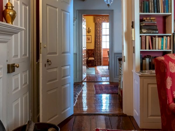 A well-furnished hallway with wooden floors, white doors, a bookshelf, and warm lighting, leading to another room.