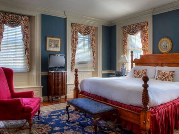 A cozy Newport, RI, guest room with a four-poster king-size bed, lamps, a red armchair, a bench, and paintings hung on the walls.