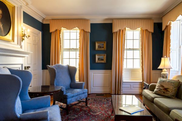 A cozy living room with blue walls, yellow curtains, blue chairs, a green couch, a portrait, and decorative frames. A lamp and a rug adorn the space.