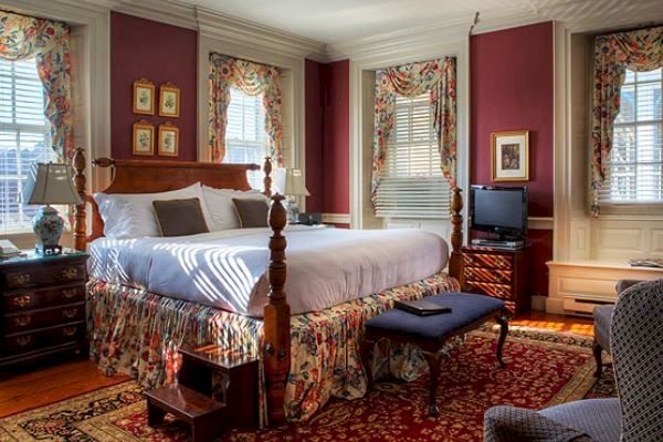 A cozy bedroom with a wooden bed, floral curtains, and matching decor. The room includes a TV, armchair, and bedside tables, featuring classic furnishings.