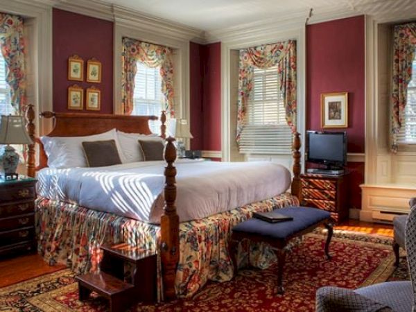 A cozy Newport, RI, guest room with a four-poster king-size bed, lamps, blue armchairs, a bench, and paintings hung on the walls.