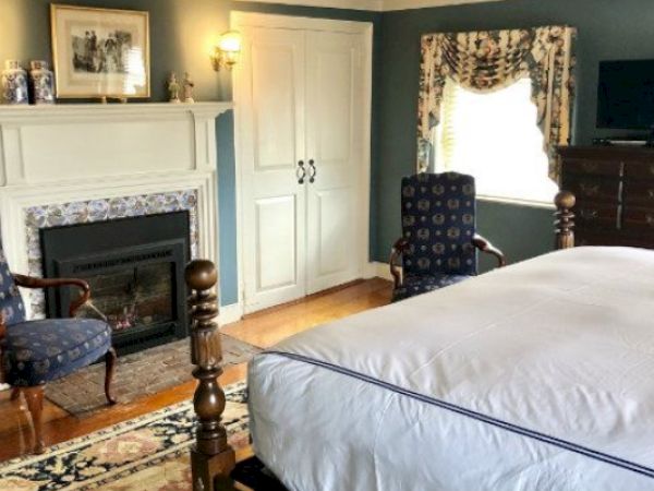 A cozy Newport, RI, guest room with a four-poster king-size bed, a gas fireplace, lamps, two blue armchairs, and a dresser.