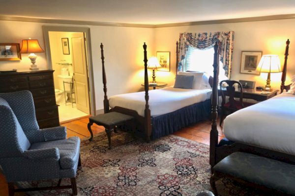 A cozy hotel room with two four-poster beds, a blue armchair, a dresser, and a desk, with a view into a well-lit bathroom.