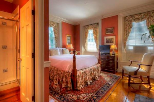 A cozy bedroom with a large bed, colorful rug, rocking chair, dresser, and window drapes, adjacent to a bathroom.