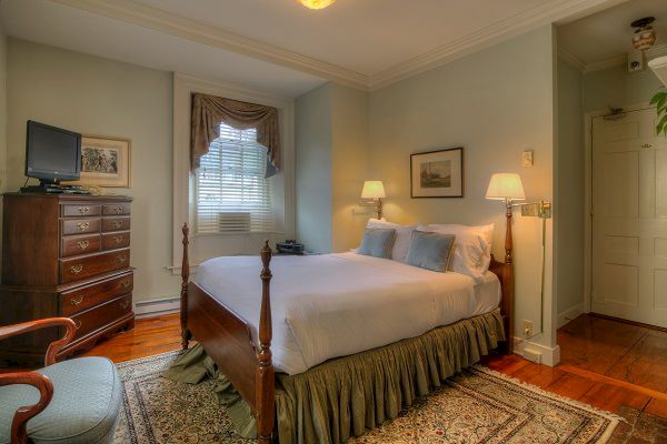 A cozy bedroom with a neatly made bed, wooden furniture, a TV on a dresser, wall art, and a window illuminating the space, ending the sentence.