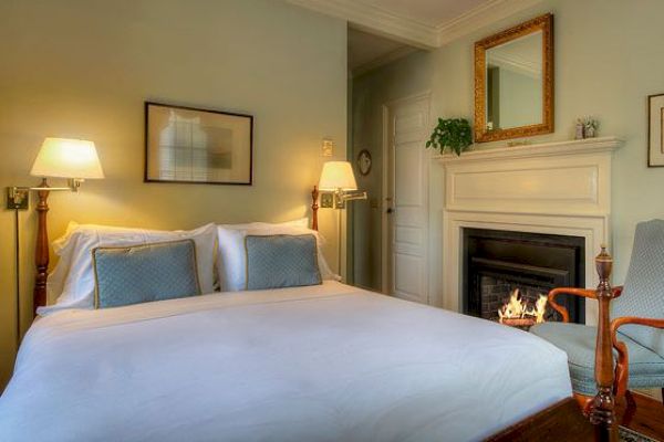 A cozy bedroom with a double bed, two wall lamps, framed artwork, a fireplace, a mirror above it, and an armchair next to the fire, ending the sentence.