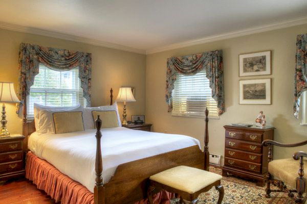 A cozy bedroom with a wooden four-poster bed, two windows with floral drapes, two bedside tables with lamps, and framed pictures on the wall.