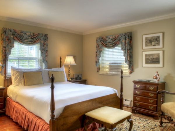 A cozy Newport, RI, guest room with a four-poster queen-size bed, lamps, a dresser, a stool, an armchair, and bright windows.