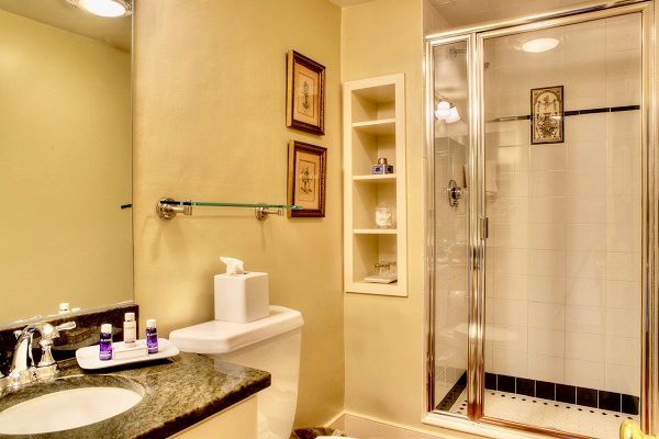 This image shows a bathroom with a glass-enclosed shower, a countertop with a sink, a toilet, and a small set of shelves. The room is warmly lit.