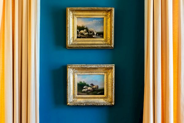 Two framed paintings are hung vertically on a blue section of a wall, flanked by beige curtains on both sides.