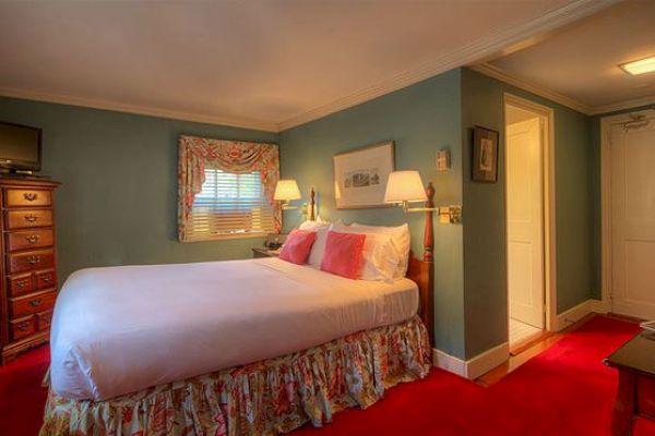 A cozy bedroom with a queen bed, floral bed skirt, two lamps, dresser with TV, artwork on walls, red carpet, and window with floral curtains.