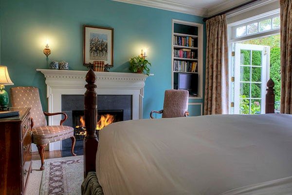 A cozy bedroom with a fireplace, chairs, a bookshelf, and a bed covered with a white sheet. There's a window showing greenery outside.