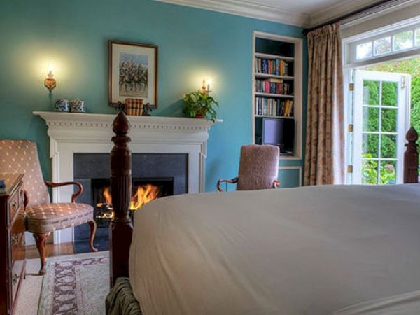 A cozy Newport, RI, guest room with a fireplace, chairs, a bookshelf, a king-size bed, and French doors leading to a garden patio.